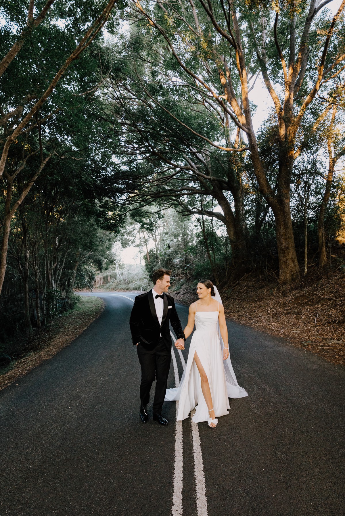 Bride and groom walk on the road with lush greenery and trees behind, photographed by wedding photographer Kirk Willcox Photography